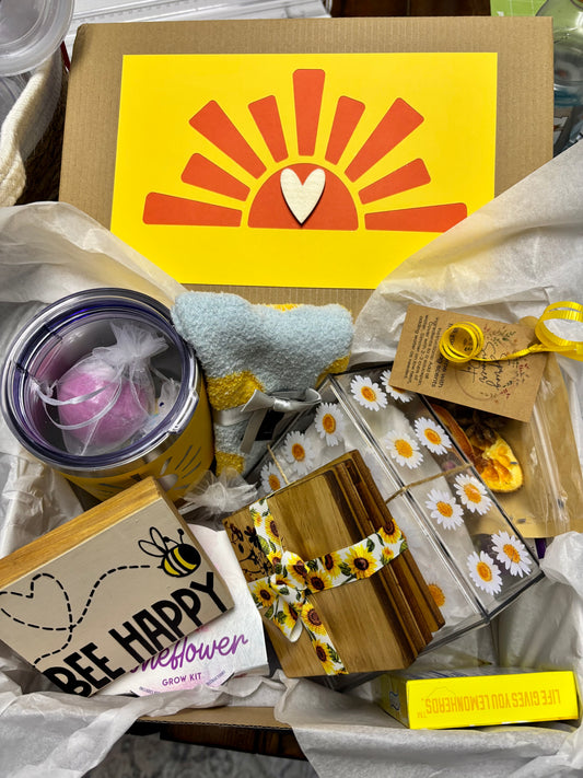 A gift box with a variety of items including a candle, a book, and other small gifts, all wrapped in decorative paper with a cheerful design featuring a sun and heart.