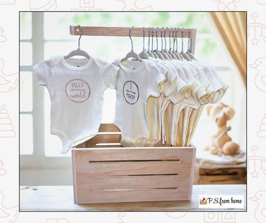 Baby onesies on hangers with wooden crate and baby shoes in a room setting.