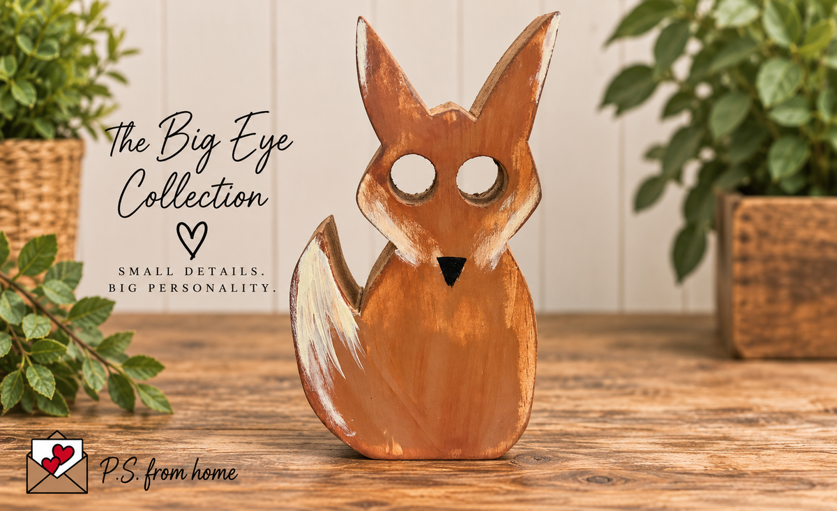 A handcrafted wooden fox figurine with large white eye cutouts, displayed on a rustic wooden table with decorative plants in the background. Text overlays read 'The Big Eye Collection' and 'PS from home'.