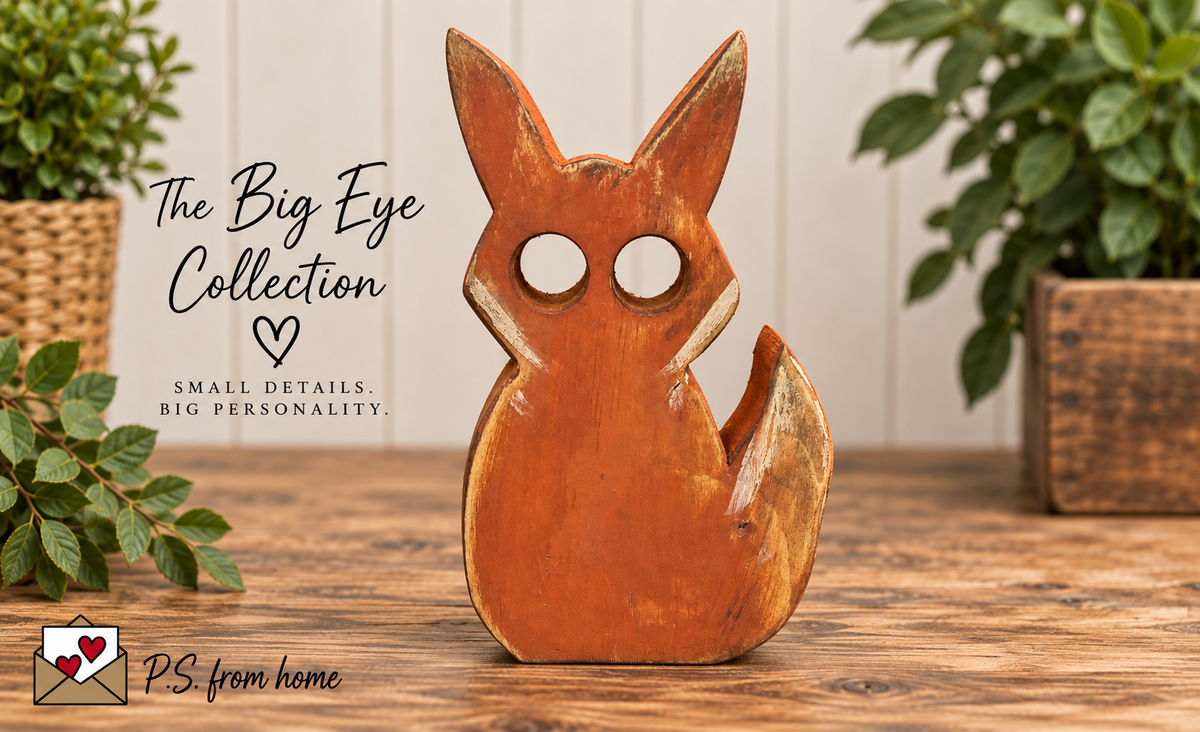 A handcrafted wooden fox figurine with large circular eye cutouts, displayed on a rustic wooden table with green plants in the background. Text on the left reads 'The Big Eye Collection' and 'Small Details.