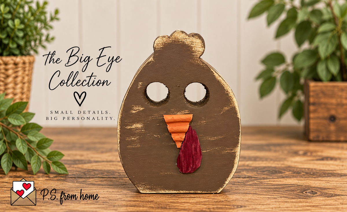 A handcrafted wooden decorative sign shaped like a chicken face, painted in distressed brown with a painted gold beak, white circular eyes, an orange triangular beak, and a red tongue. It is displayed on a wooden table with a blurred background of potted plants and a sign reading 'the Big Eye Collection'.