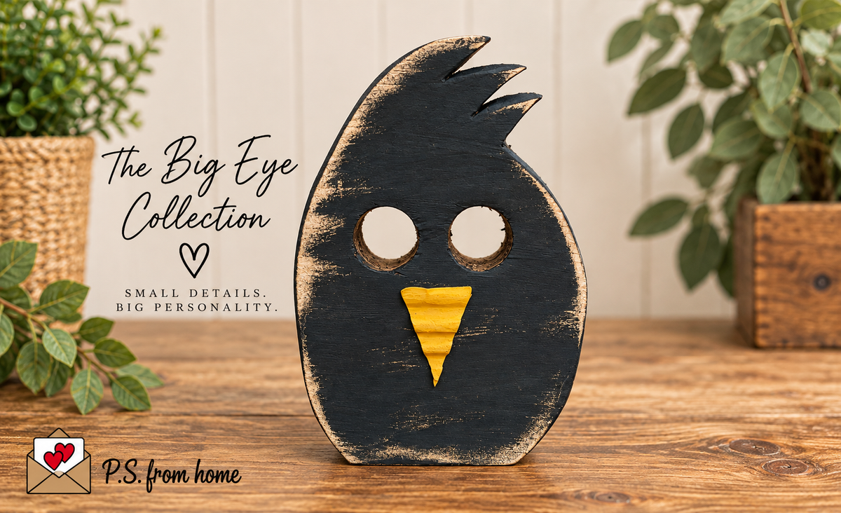 A black wooden decorative bird figure with a large oval face, two circular eye cutouts, and a yellow beak, standing on a wooden surface. The image includes the text 'The Big Eye Collection' and 'SMALL DETAILS.
