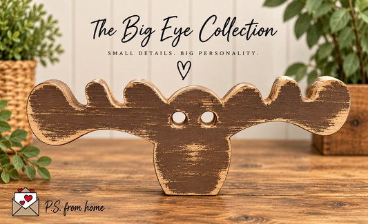 A rustic wooden moose head sign with distressed brown and tan paint, displayed on a wooden surface with green plants in the background. Text at the top reads 'The Big Eye Collection' with smaller tagline 'SMALL DETAILS.