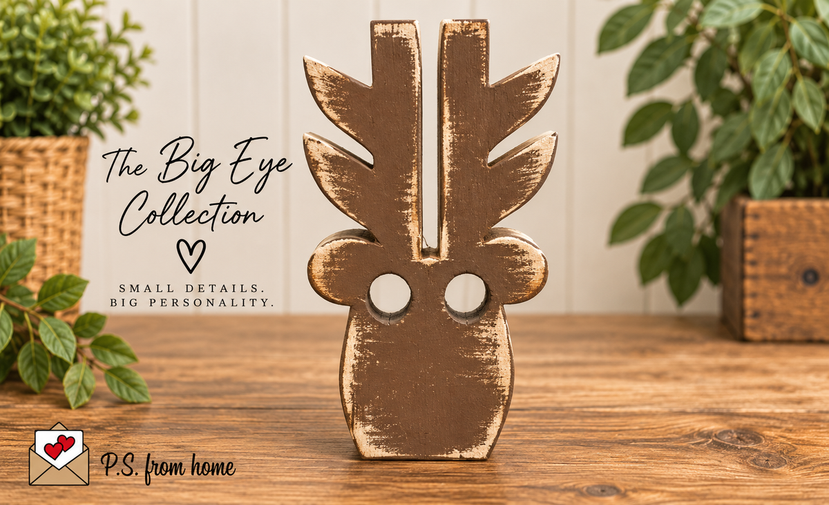 A rustic, distressed brown wooden decorative piece shaped like a stylized face with large circular cutouts, standing on a wooden tabletop. The background features a white paneled wall, potted green plants, and a text overlay reading 'The Big Eye Collection' with a 'P.