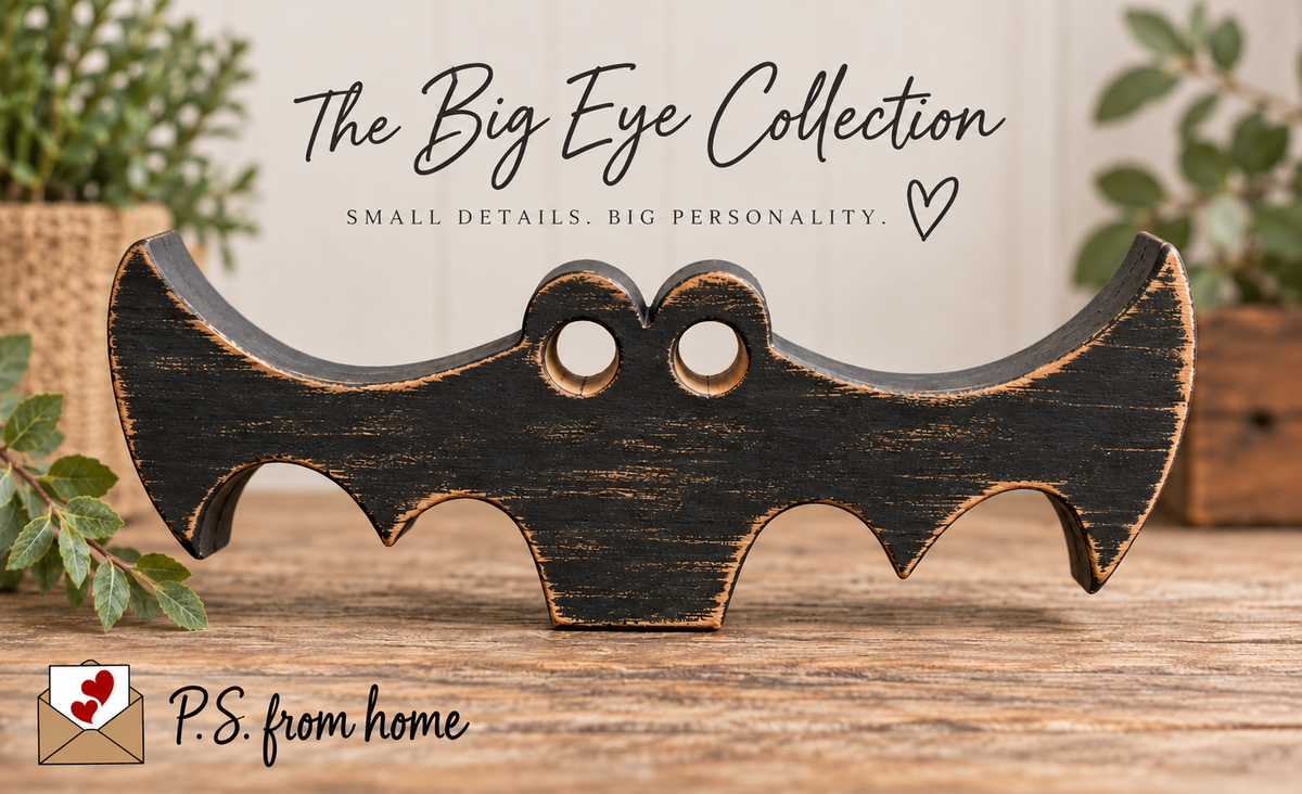 A distressed black wooden bat-shaped wall decor piece with a rustic brown wood-grain finish, displayed on a wooden surface with decorative plants in the background. Text overlay at the top reads 'The Big Eye Collection'.