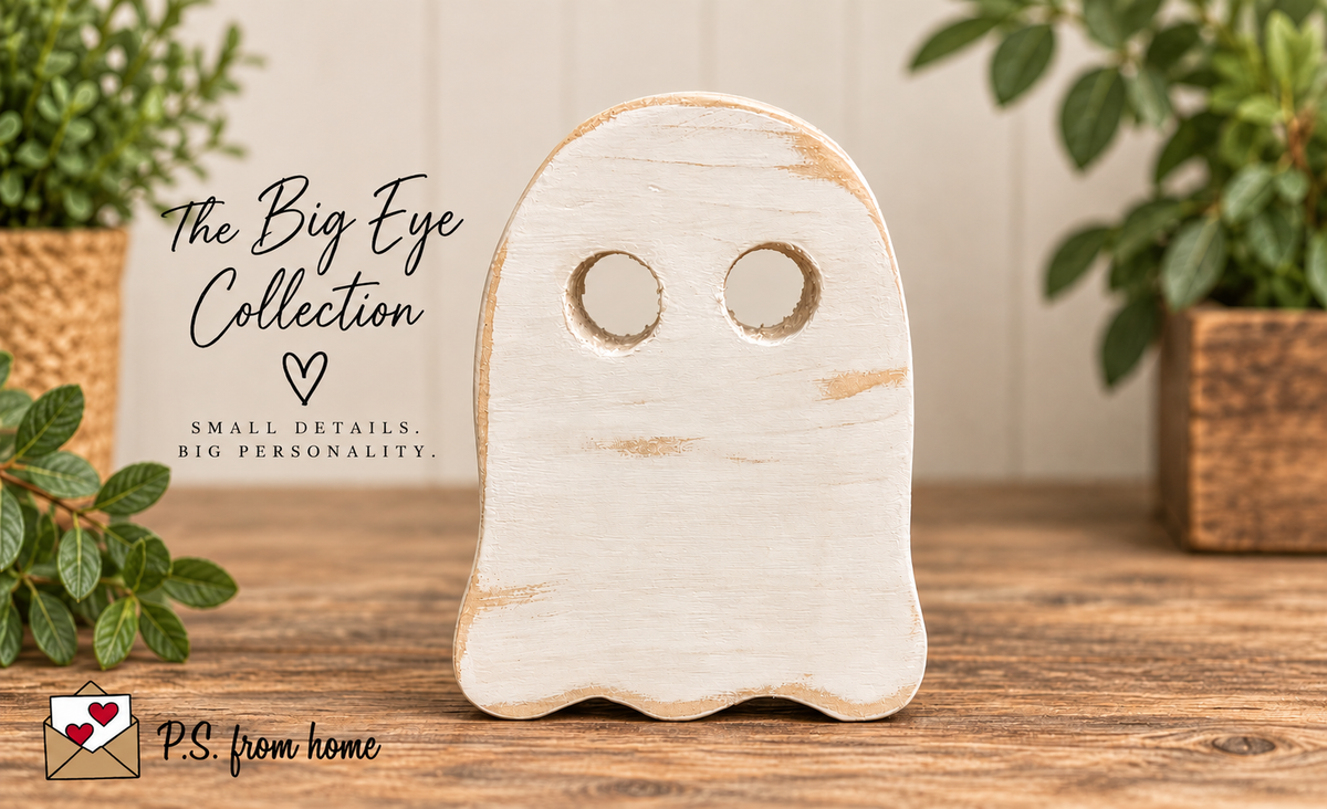 A ghost-shaped wooden decor piece with two circular eye cutouts, standing on a wooden surface. The product is shown in a styled home setting with potted plants and a sign reading 'The Big Eye Collection' and 'P.
