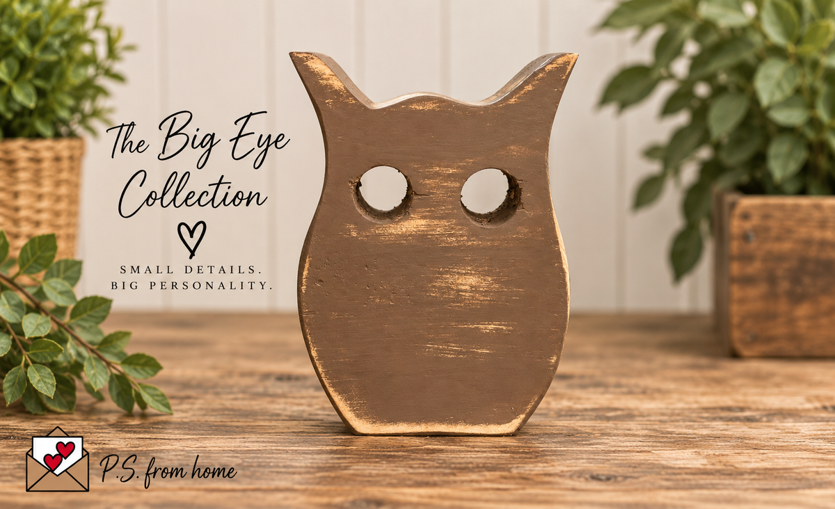 A rustic wooden owl figurine with distressed brown and gold paint, featuring large circular cutouts for eyes, displayed on a wooden table with decorative plants in the background. Text overlays read 'The Big Eye Collection' and 'PS from home'.