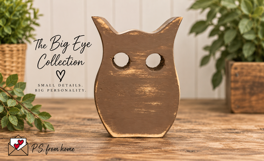 A rustic wooden owl figurine with distressed brown and gold paint, featuring large circular cutouts for eyes, displayed on a wooden table with decorative plants in the background. Text overlays read 'The Big Eye Collection' and 'PS from home'.