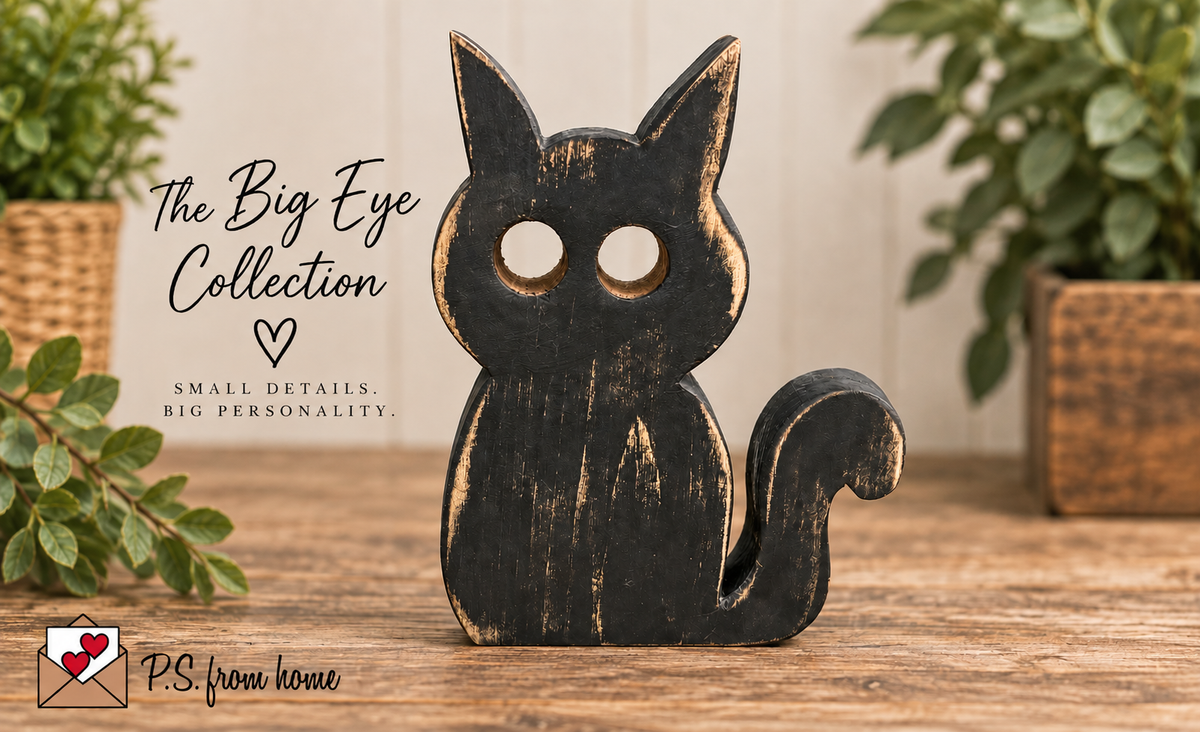 A black wooden cat figurine with a distressed finish and large eye cutouts, displayed on a wooden surface with decorative plants in the background. Text on the left reads 'The Big Eye Collection' and 'PS from home'.