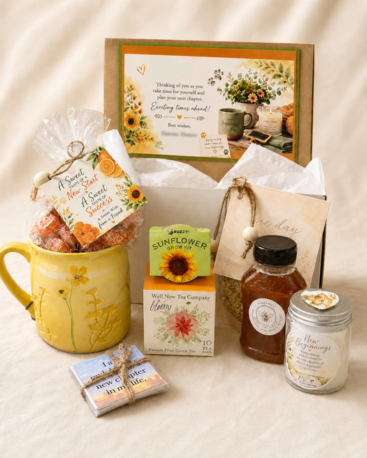 A curated gift basket arrangement on a cream fabric background, featuring a yellow ceramic mug filled with sunflower candies, a framed letter, a box of Muffin Man Tea Company tea, a jar of honey, a candle, and small booklets.