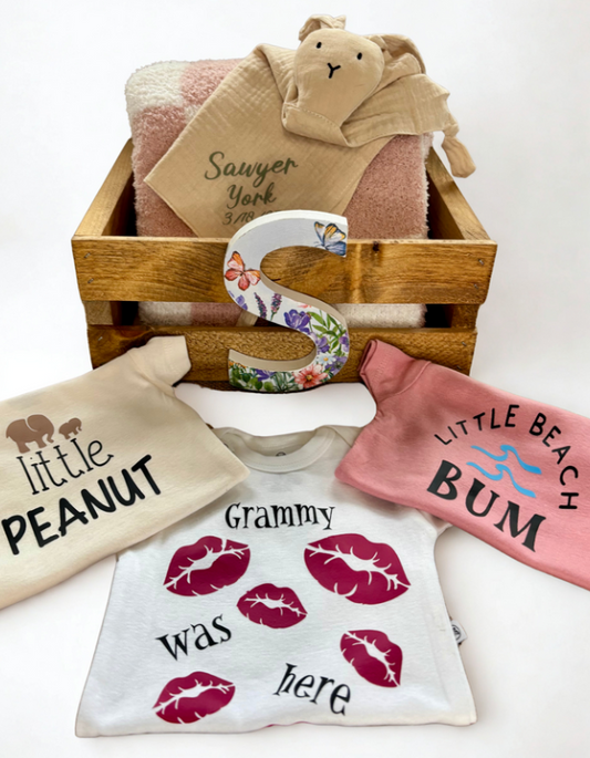 Collection of baby-themed items including t-shirts, a blanket, and a decorative letter 'S' in a wooden crate.